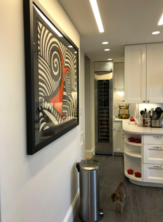 Modern kitchen with white cabinets and a stainless steel wine cooler. A colorful abstract painting hangs on the wall, and a cat stands on the floor near a silver trash can.