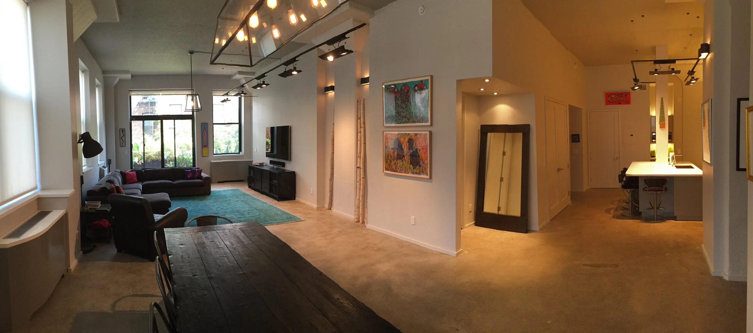 An open-concept living room and kitchen area with large windows, a dark sectional sofa with red and purple pillows, a television on a black stand, artwork on the walls, and a bar counter with stools.