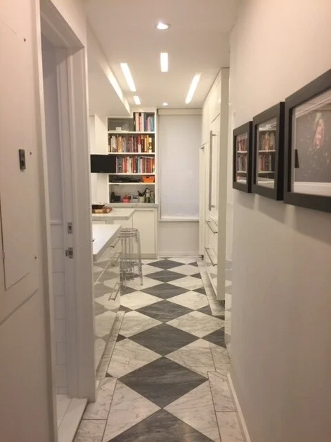 A hallway with black and white checkered marble floor, white walls, and framed photos on the right wall. At the end, a bookshelf filled with colorful books and a small kitchenette with a counter and clear stools.