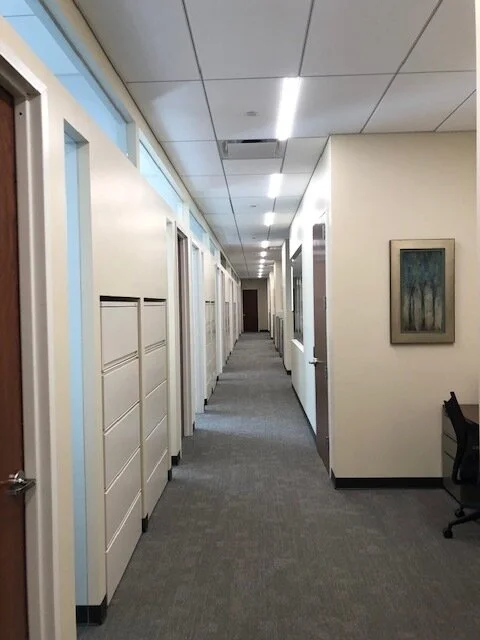 Empty office corridor with closed doors on the left, a chair on the right, and framed artwork on the wall.