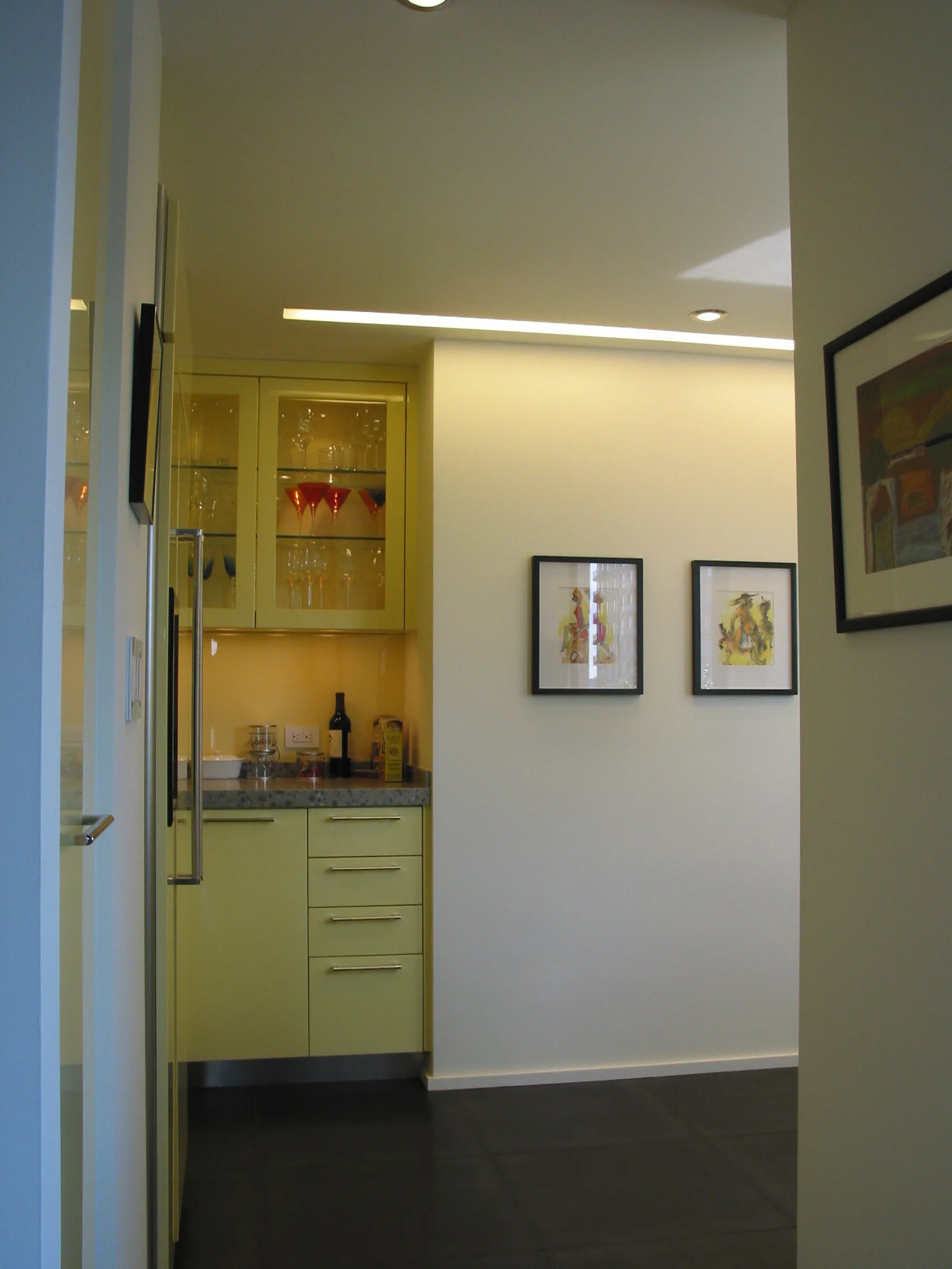 View of a small kitchenette and adjacent wall with framed artwork, yellow cabinetry, and glassware in a modern apartment.