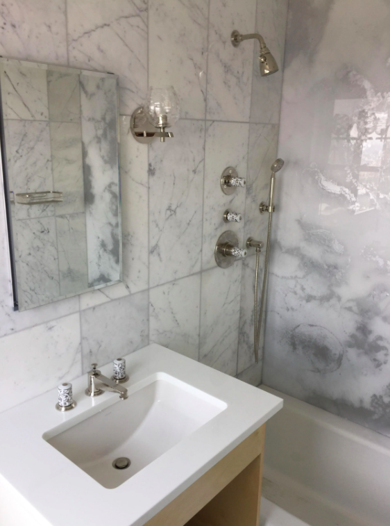 Bathroom with marble walls, a white sink with three chrome faucet handles, a mirror, and a shower area with multiple shower controls and a handheld showerhead.
