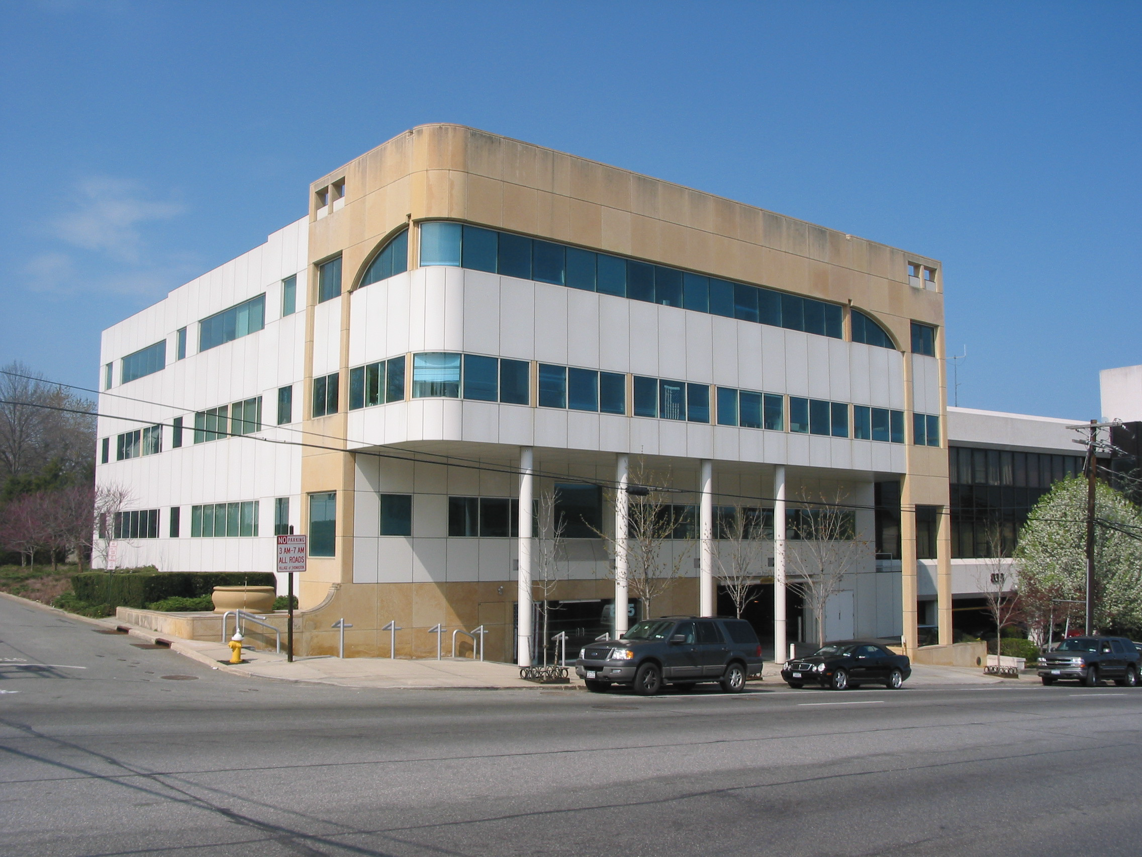 A modern multi-story office building with white and tan panels, green-tinted windows, and supporting columns at the entrance. Cars are parked in front and a street runs past the building, with a bright blue sky overhead.