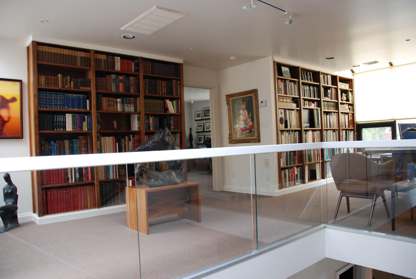 A cozy library room with built-in bookshelves filled with books, a framed painting of a young girl on the wall, a sculpture of a face on a small wooden table, and a glass railing separating the space from an area below.