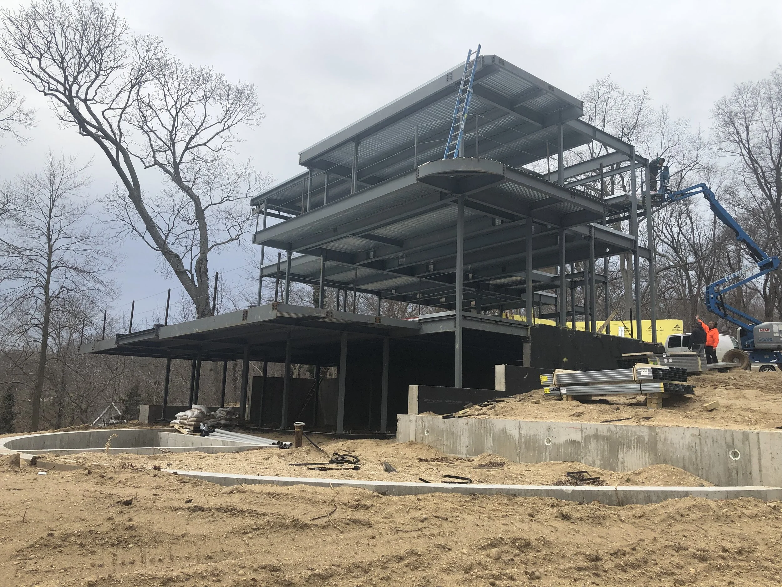 Construction site with a multi-story steel frame structure being built, workers in safety gear, a crane lift, and leafless trees in the background.