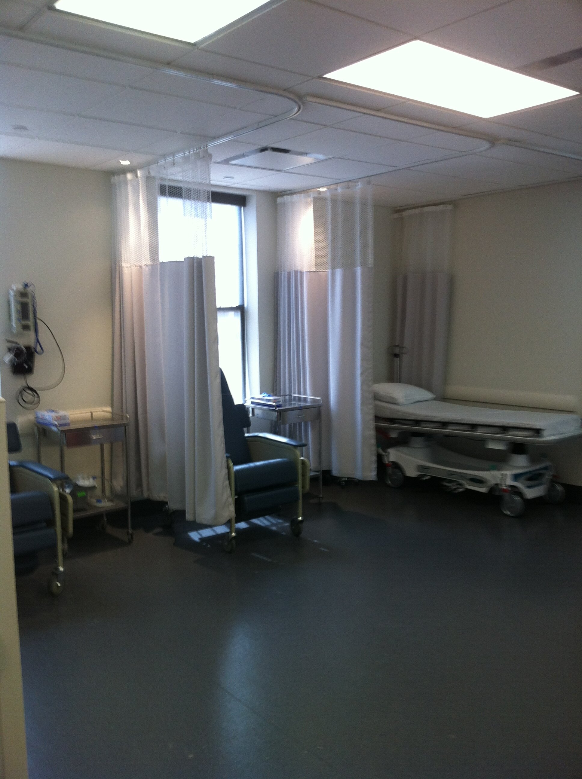 A hospital room with a bed, medical equipment, a green chair, and curtains separating the bed from the rest of the room. The room is well-lit with overhead fluorescent lights and has a window with curtains.