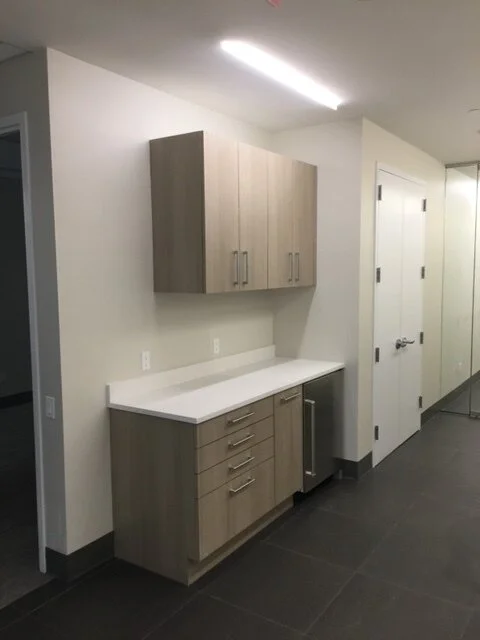 A kitchenette with light wood cabinets, a white countertop, and a small refrigerator, in a modern, minimalist style.
