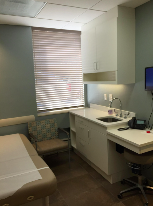 Medical examination room with a window covered by blinds, a patient bed, a chair, cabinets, a sink, a telephone, and a small wall-mounted monitor.