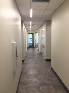 An empty hallway in a modern building with white walls, gray flooring, ceiling lights, and a window at the end.