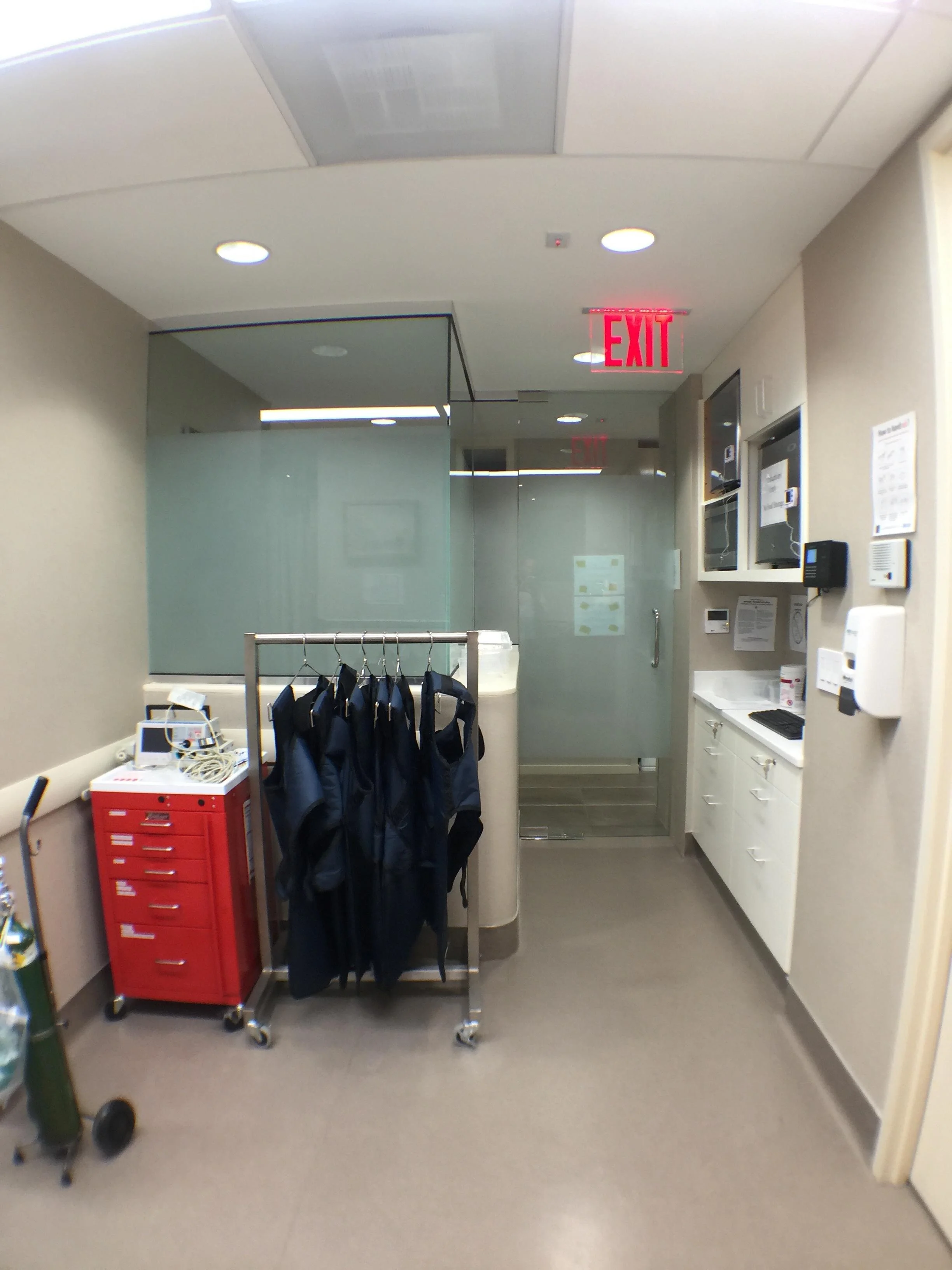 Hospital corridor with a red exit sign, medical uniforms hanging on a rack, medical equipment, and cabinets with supplies.