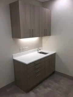 A small kitchen area with wooden cabinets, a white countertop, and a sink, against plain white walls.