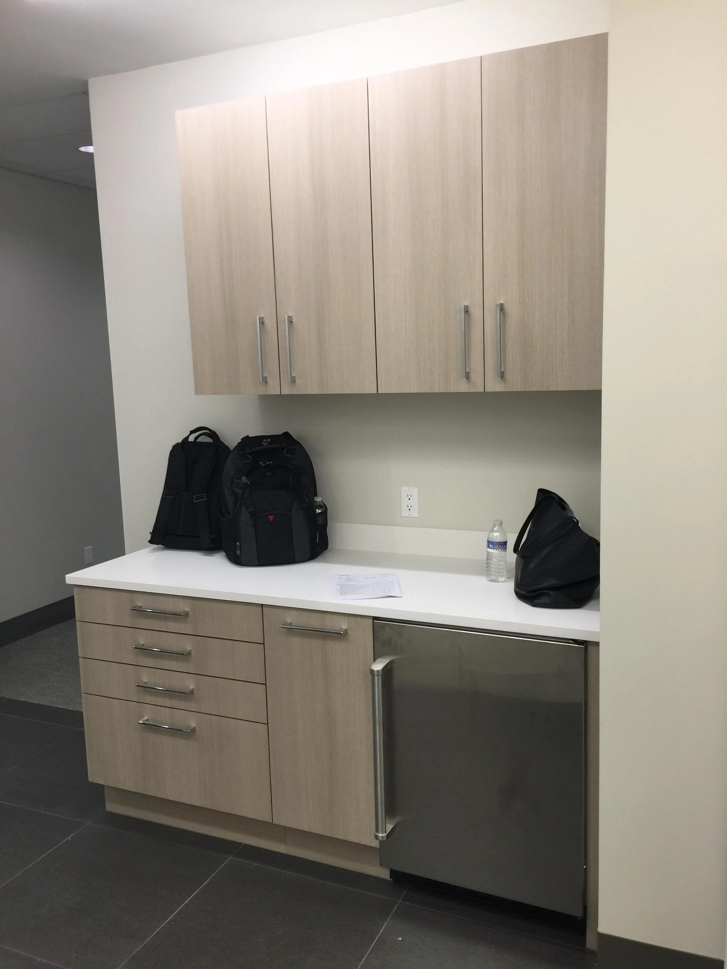 A kitchenette with beige cabinets, a white countertop, a black water bottle, a black bag, a backpack, and a piece of paper on the counter. There is also a small refrigerator under the counter and an electrical outlet on the wall.