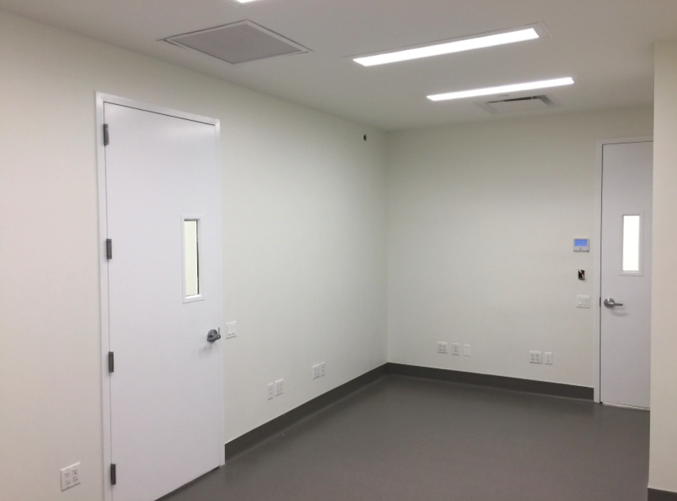 Empty, white-walled room with fluorescent ceiling lights, gray flooring, two doors with small windows, and multiple electrical outlets.