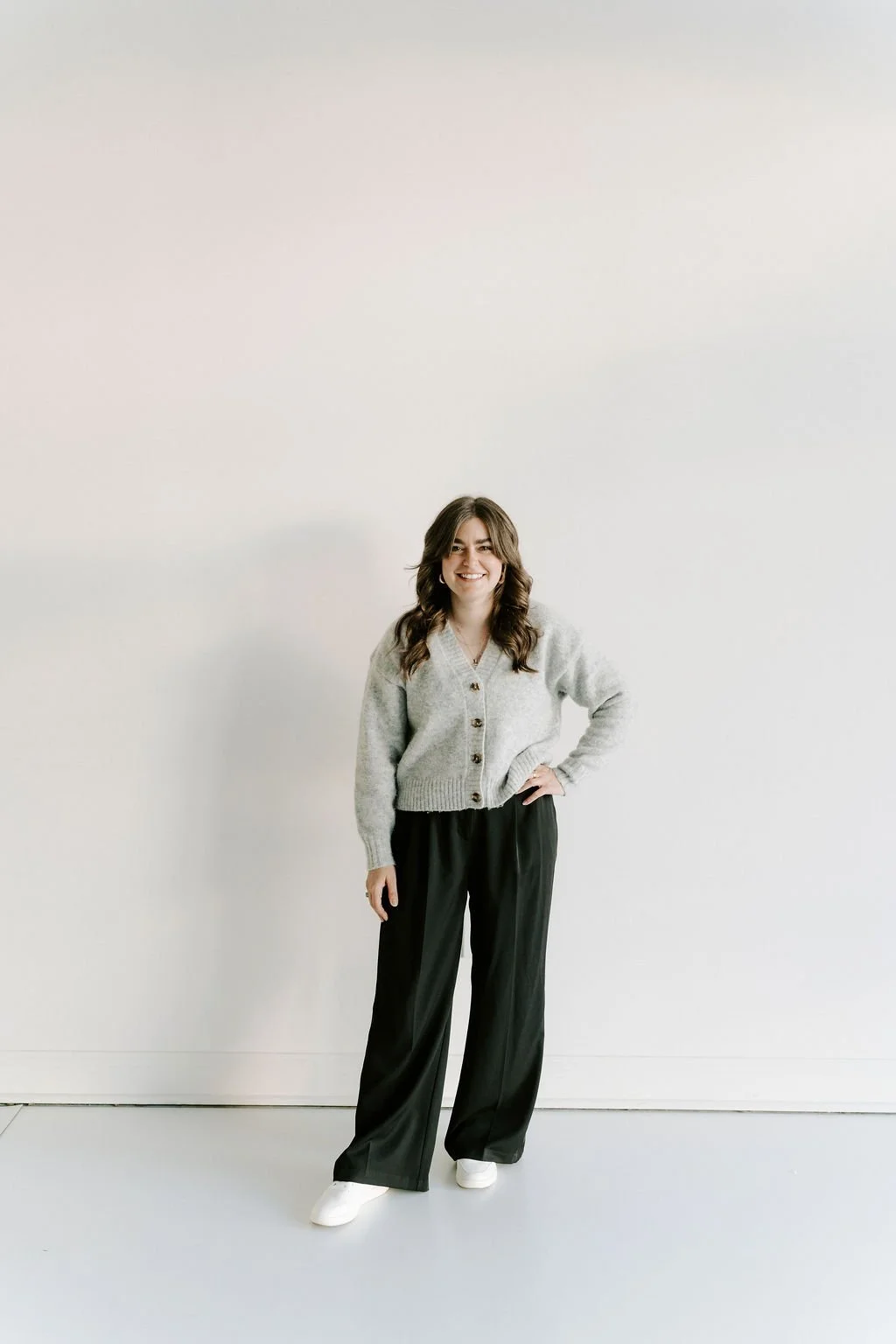 A woman with long brown hair, smiling, wearing a light gray cardigan, black pants, and white shoes, standing against a plain white wall.