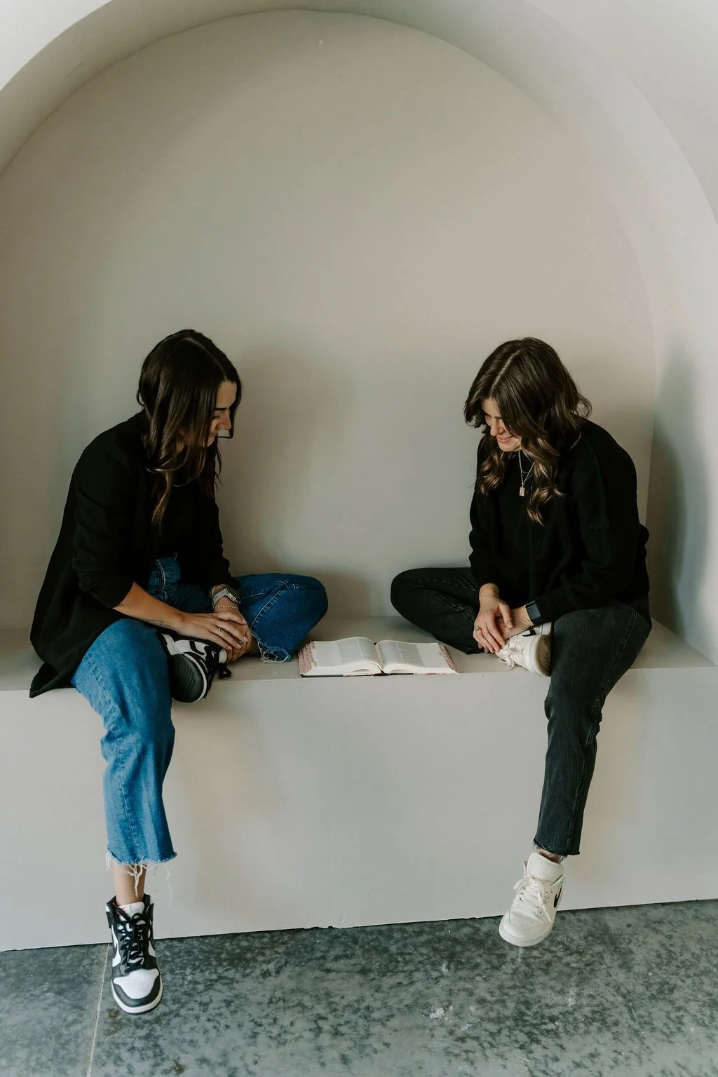Two women sitting on a white ledge, looking at an open book with a plain white wall background.