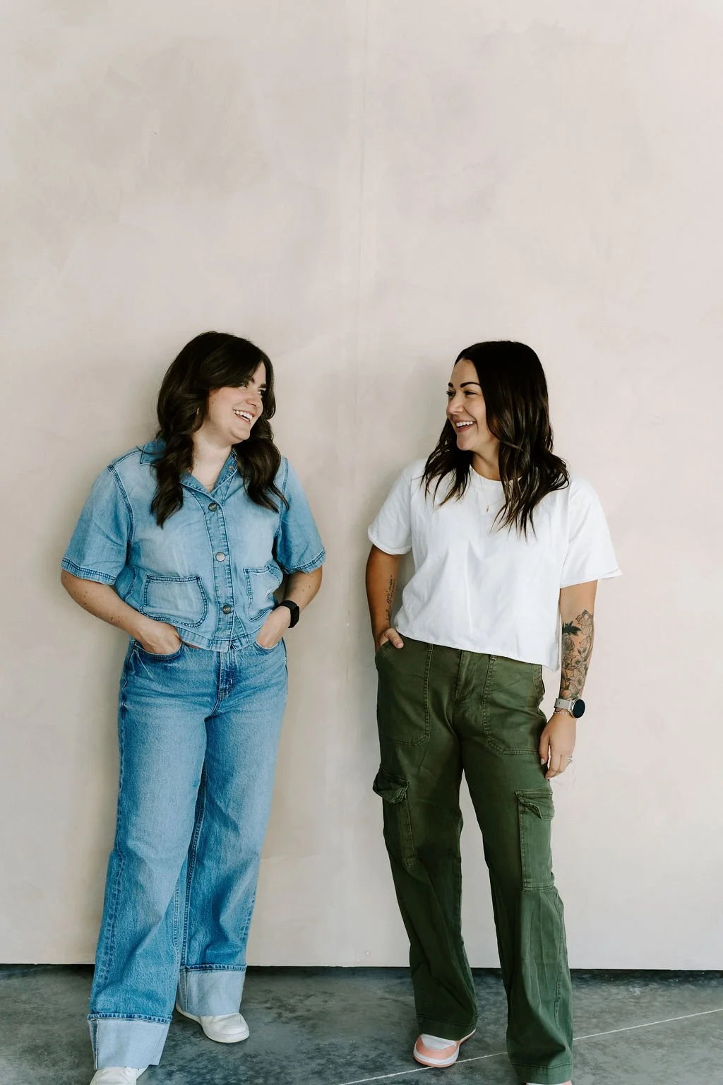 Two women standing against a plain beige wall, smiling and laughing while looking at each other.