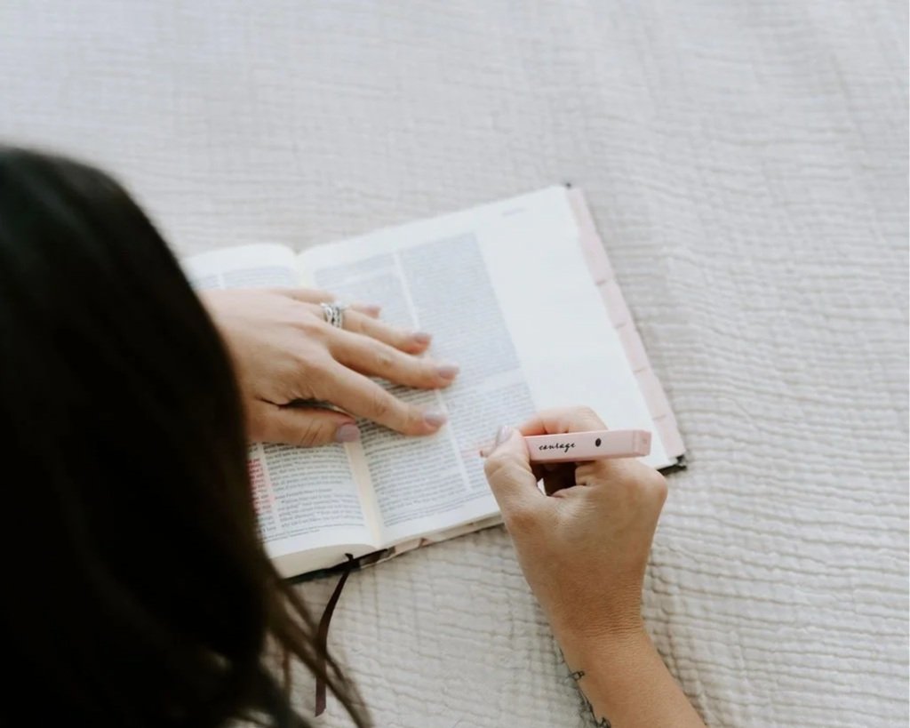 A person reading a book on a textured surface, writing in a planner with a pink pen.