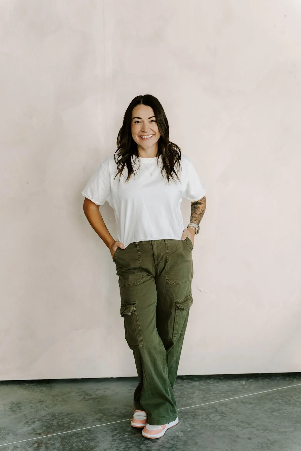 A woman with dark brown hair and tattoos on her left arm, smiling, standing against a plain light-colored wall, wearing a white T-shirt, baggy green cargo pants, and white sneakers.