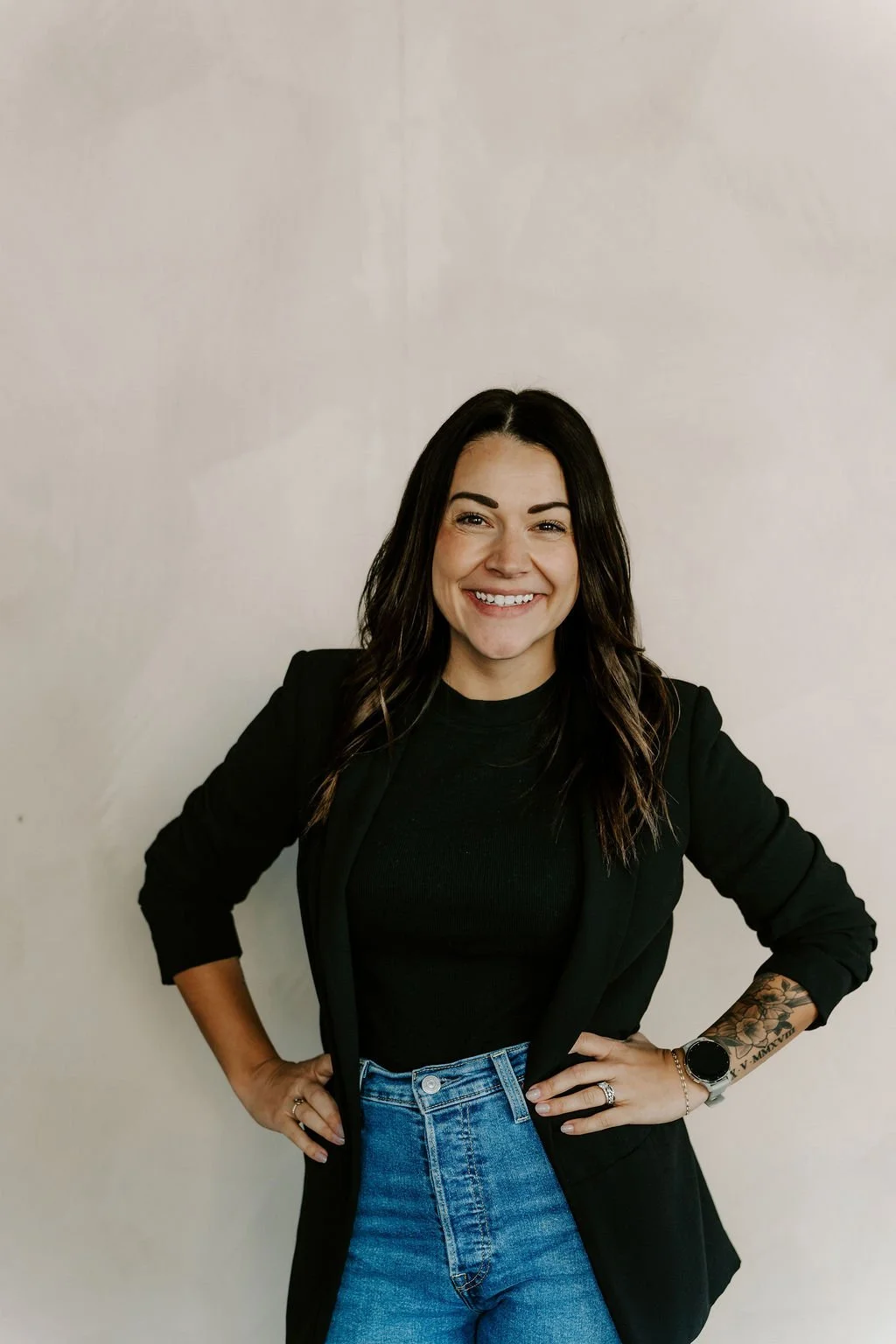 Woman with dark hair smiling, wearing a black blazer, black top, and blue jeans, standing against a plain light-colored background.