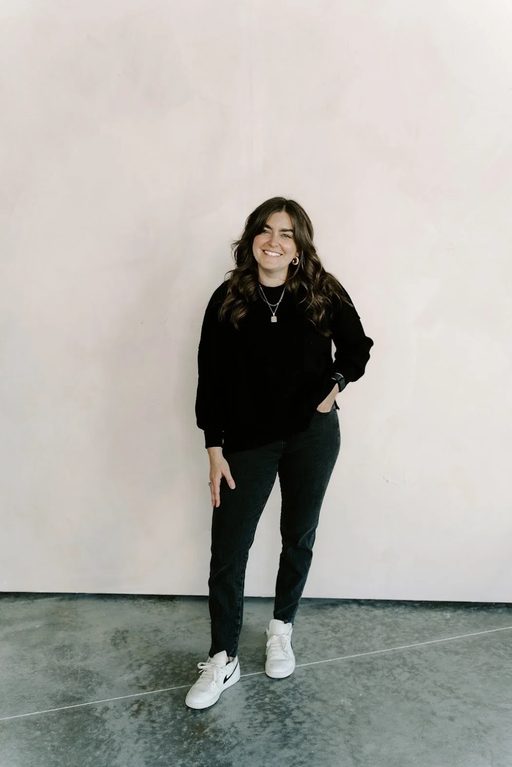 A woman standing against a plain wall, wearing a black sweatshirt, black pants, and white sneakers, smiling with one hand in her pocket.