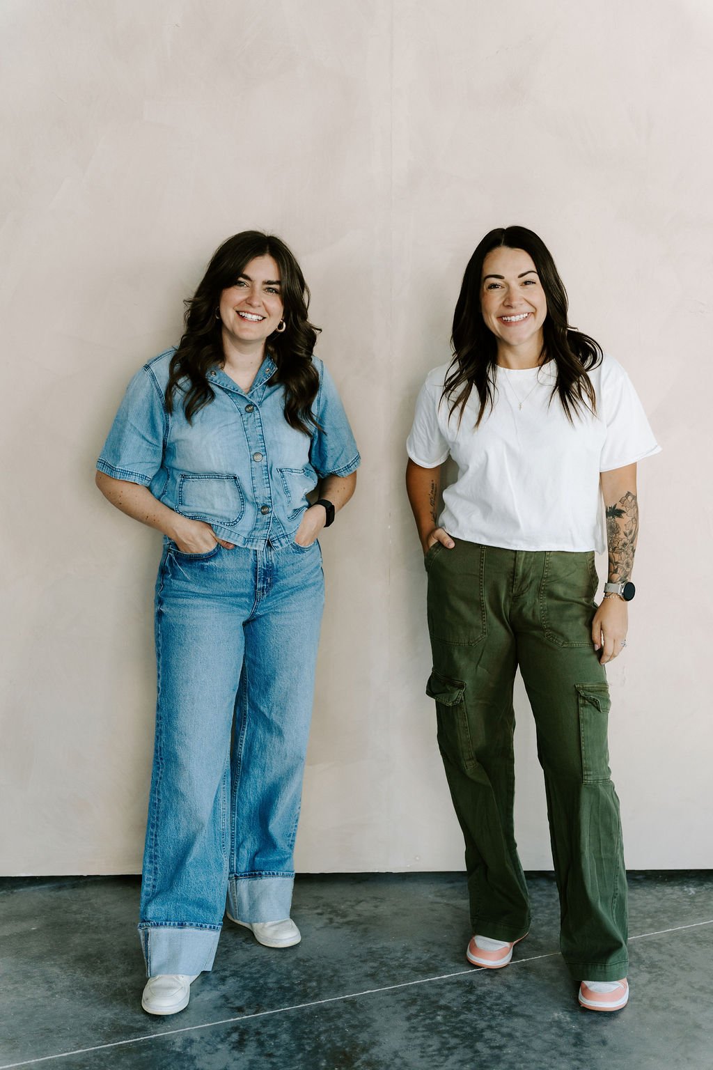 Two women standing side by side against a plain beige wall, smiling at the camera. The woman on the left is wearing a light blue denim jacket and jeans, while the woman on the right is wearing a white t-shirt and olive green cargo pants.