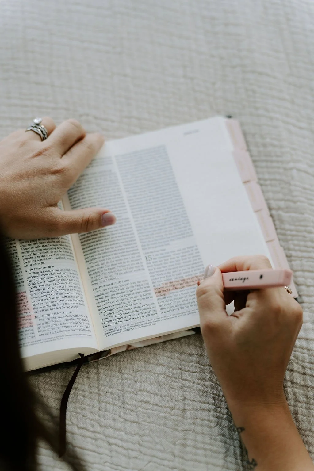 A person holding a pink highlighter in the right hand and turning the page of a Bible with the left hand, while sitting on a cream-colored textured surface.