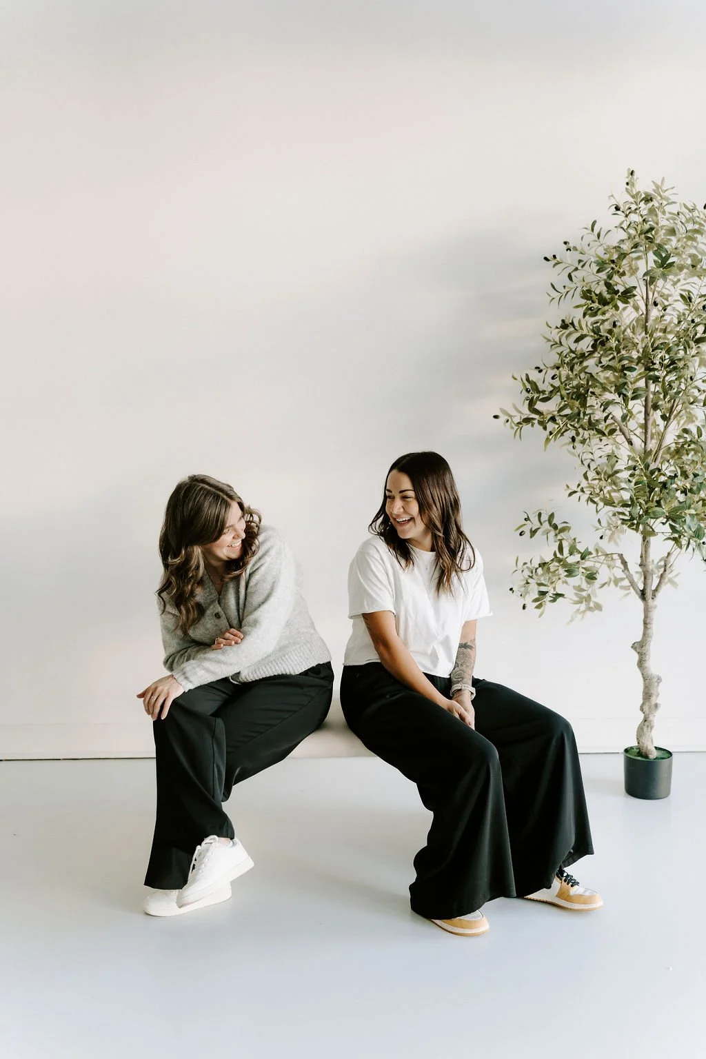Two women sitting on a white bench, smiling and looking at each other, with a potted tree on the right side of the image, against a plain white background.