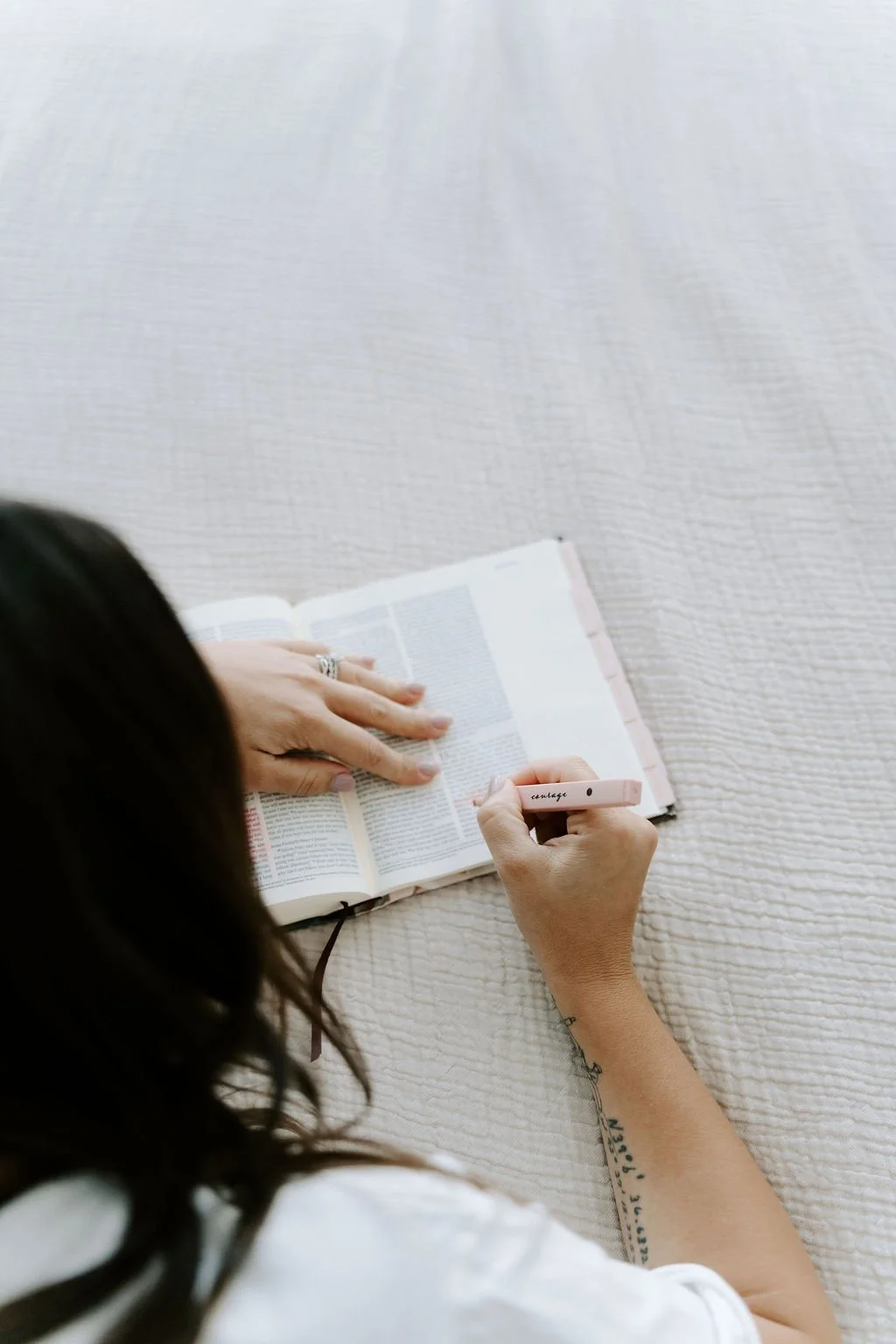 Person reading a Bible on a cream-colored textured surface while writing in it with a pink highlighter.