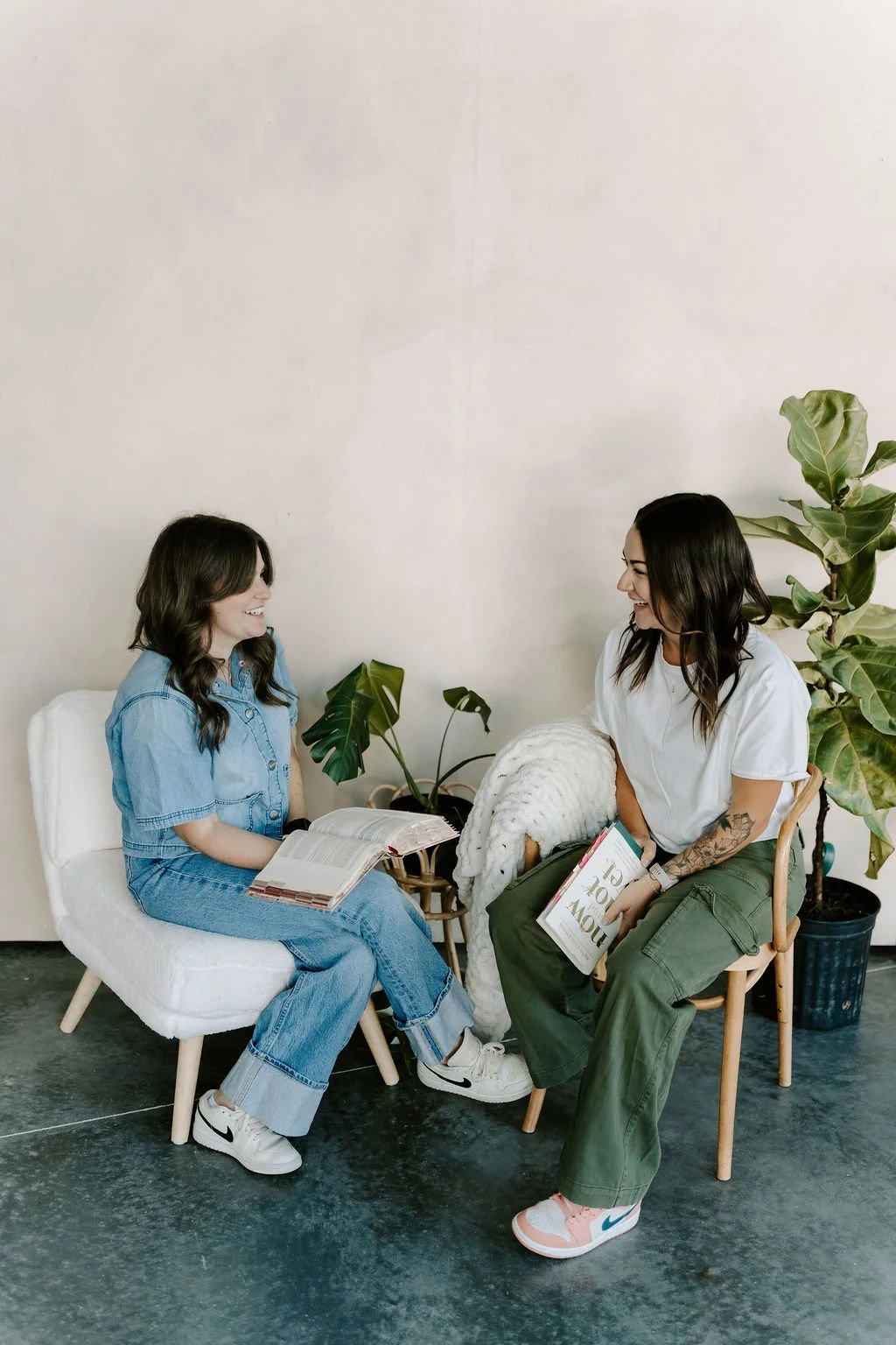 Two women sitting and smiling at each other in a cozy cafe, exchanging books, with potted plants and neutral decor behind them.