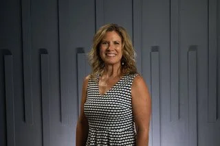 A woman with shoulder-length blonde hair smiling, wearing a sleeveless black and white patterned dress, standing in front of a light gray background with vertical panels.