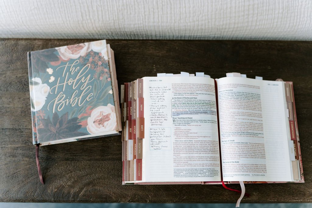 Open Bible with tabs and handwritten notes, next to a closed Bible titled 'the Holy Bible' with floral cover on a wooden surface.