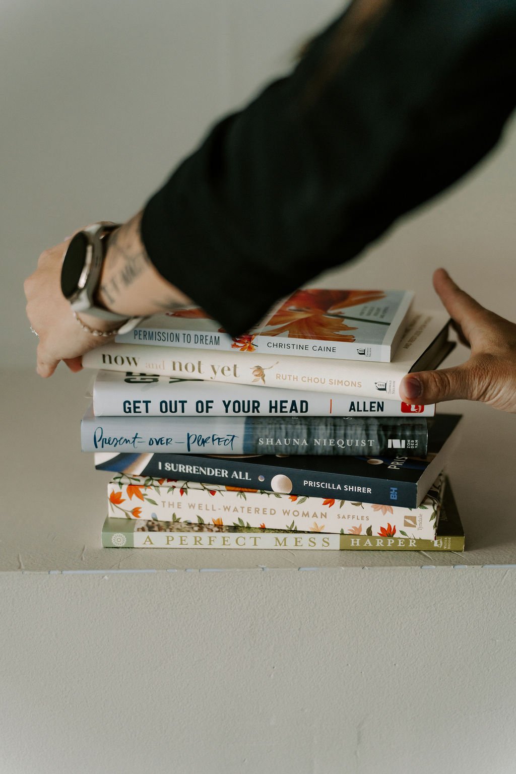Stack of seven books on a beige surface, with a person reaching to pick a book, wearing a black top and a wristwatch.