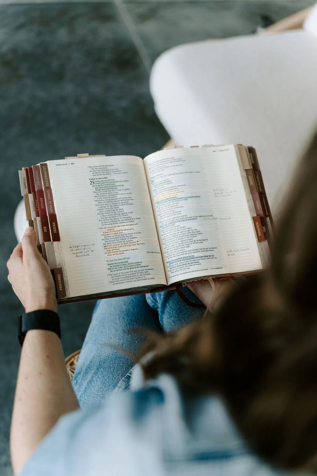 A person sitting on a gray floor, holding an open Bible with highlighted verses and handwritten notes, wearing a black wristwatch and blue jeans, with a white chair in the background.