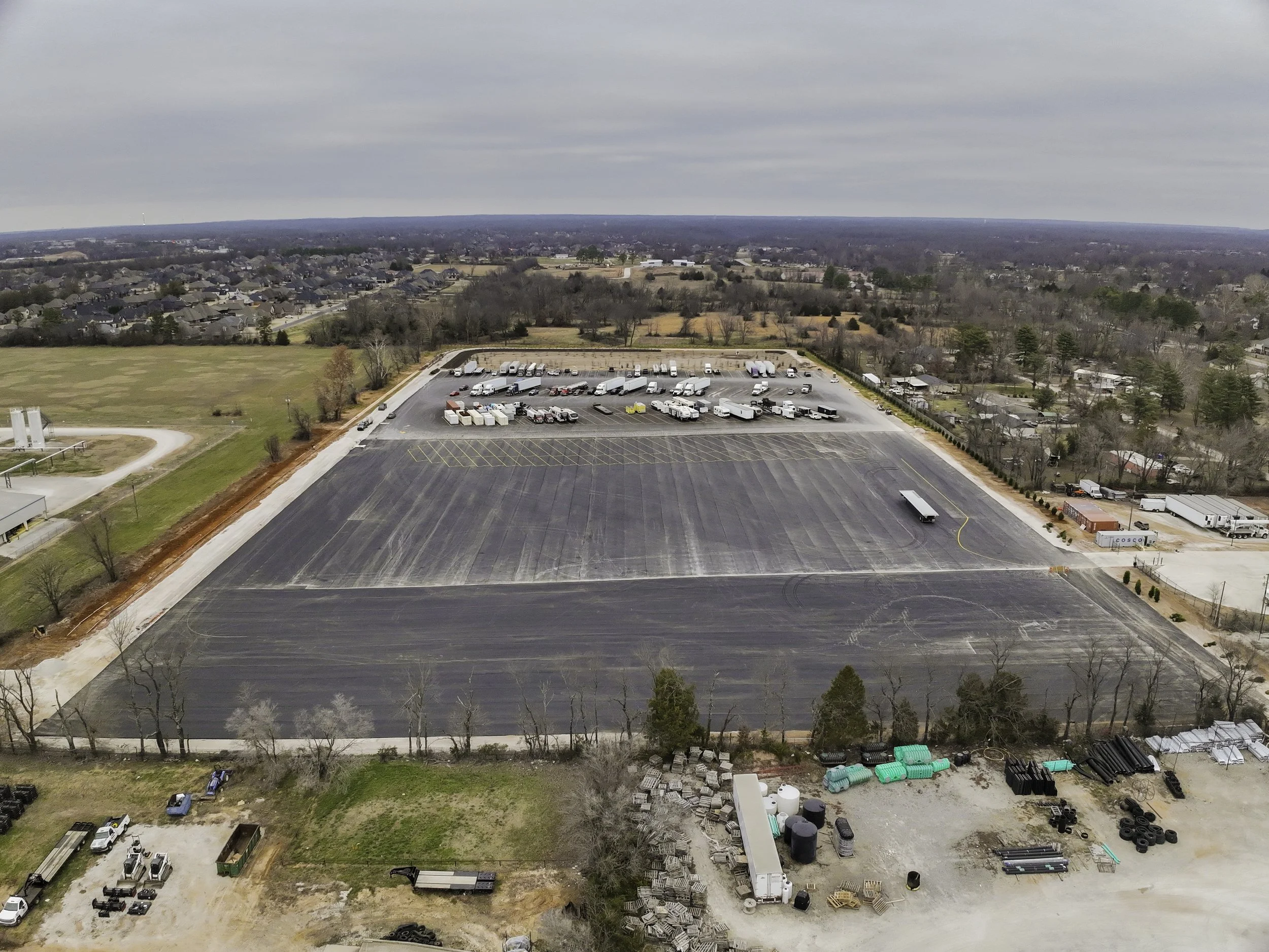 Aerial view of an empty parking lot with some parked trucks and trailers, surrounded by trees, open fields, and residential neighborhoods on a cloudy day.