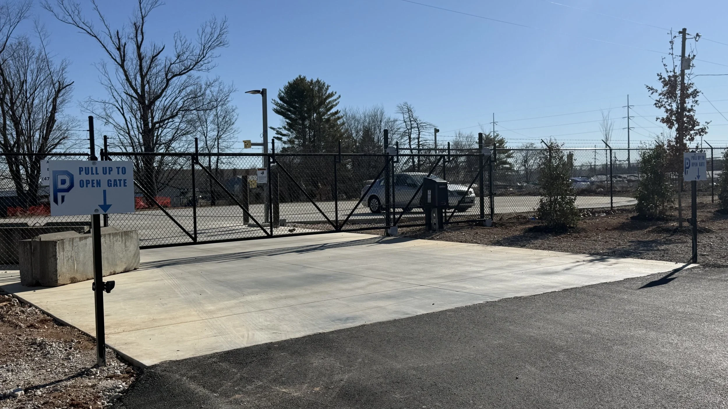 A parking lot enclosed by a chain-link fence with a gate. Signs instruct to pull up to open the gate. There are trees and power lines in the background, and a clear blue sky above.