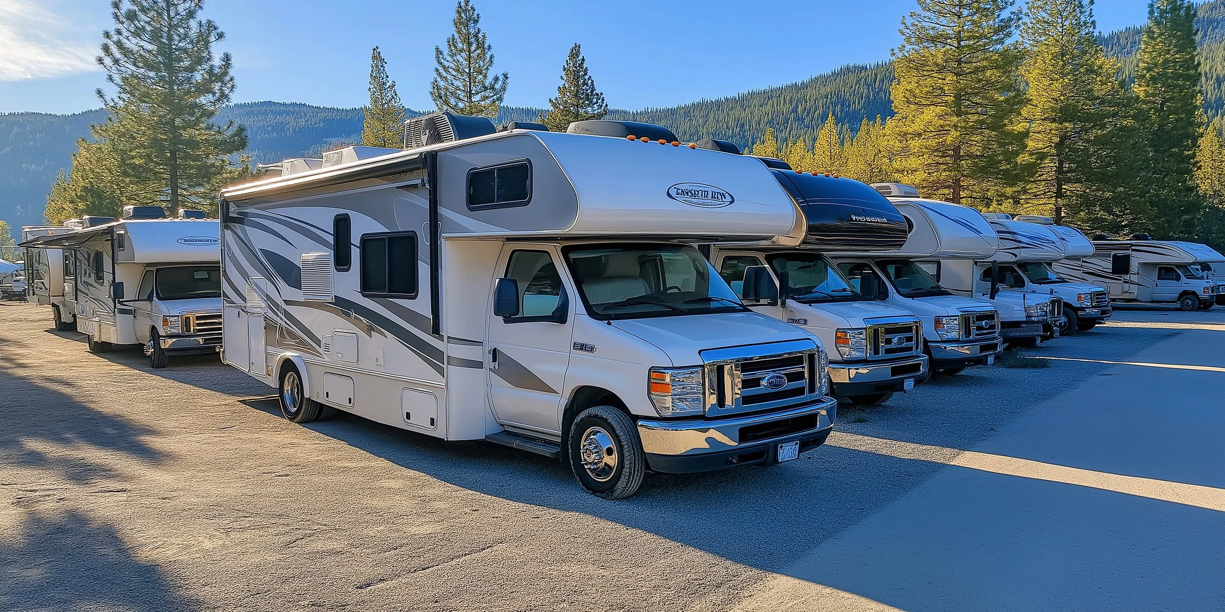 Row of recreational vehicles parked on a dirt lot with trees and mountains in the background, under a clear blue sky.