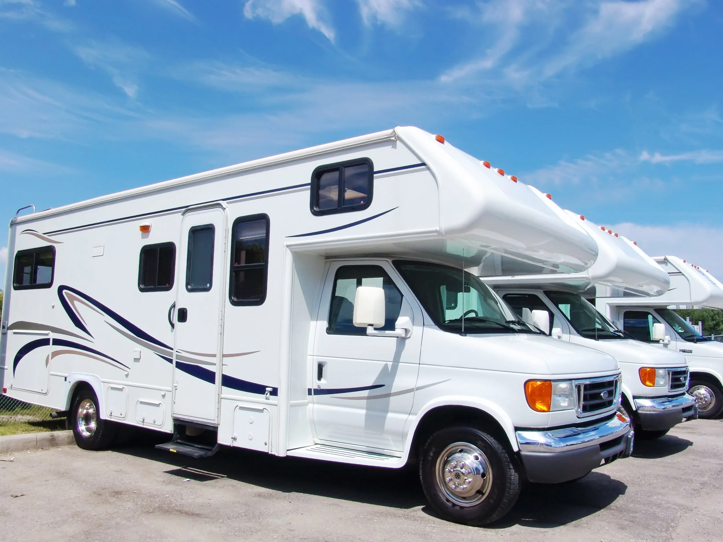 White recreational vehicle (RV) with a large over-cab sleeping area, parked on a paved lot under a blue sky with clouds.