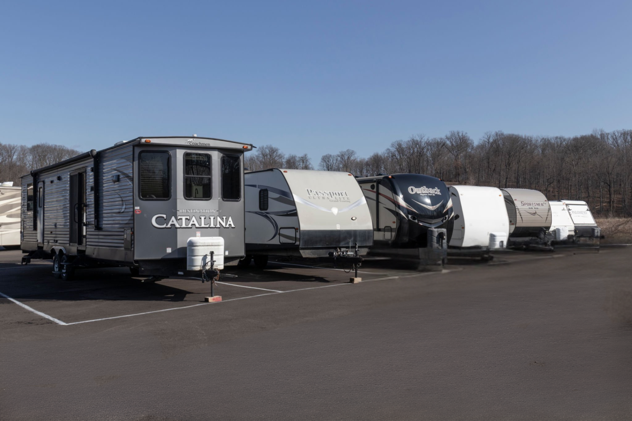 Multiple travel trailers parked side by side on a paved lot against a backdrop of leafless trees and a clear blue sky.