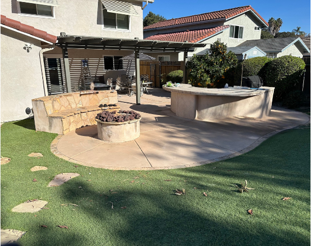 Backyard patio with stone and concrete features, a fire pit, outdoor seating, and a covered area with a roof, surrounded by shrubs and a lawn.
