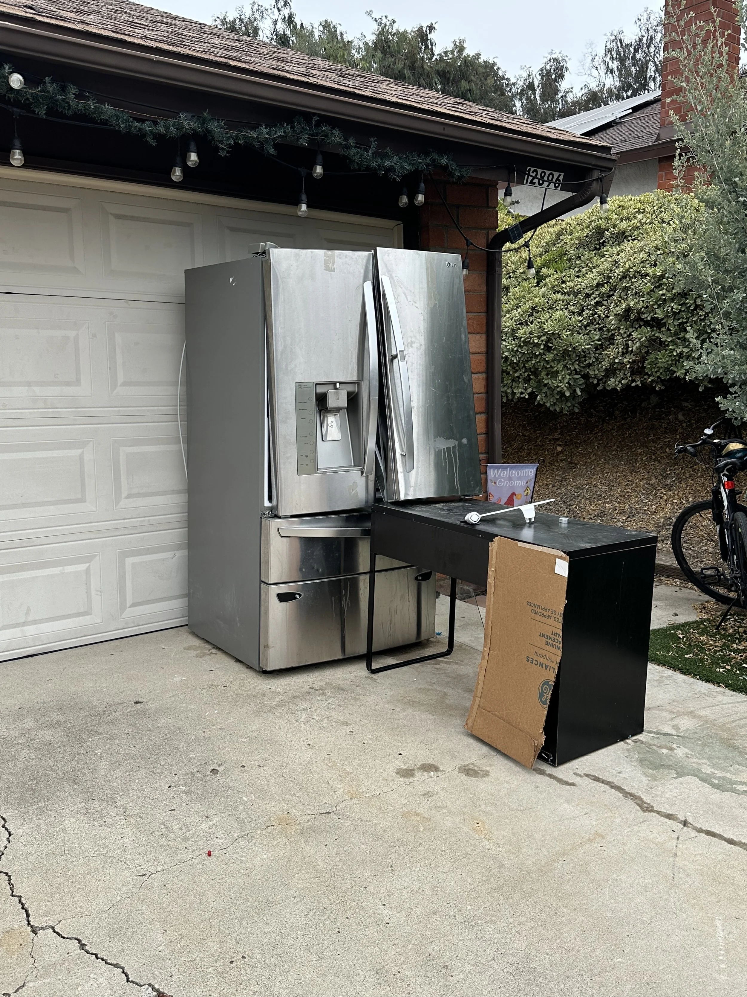 An outdoor scene with a stainless steel refrigerator placed on a concrete driveway next to a black desk, with a cardboard box leaning against it. A garage door and part of a brick house are visible, along with greenery and a bicycle on the right side