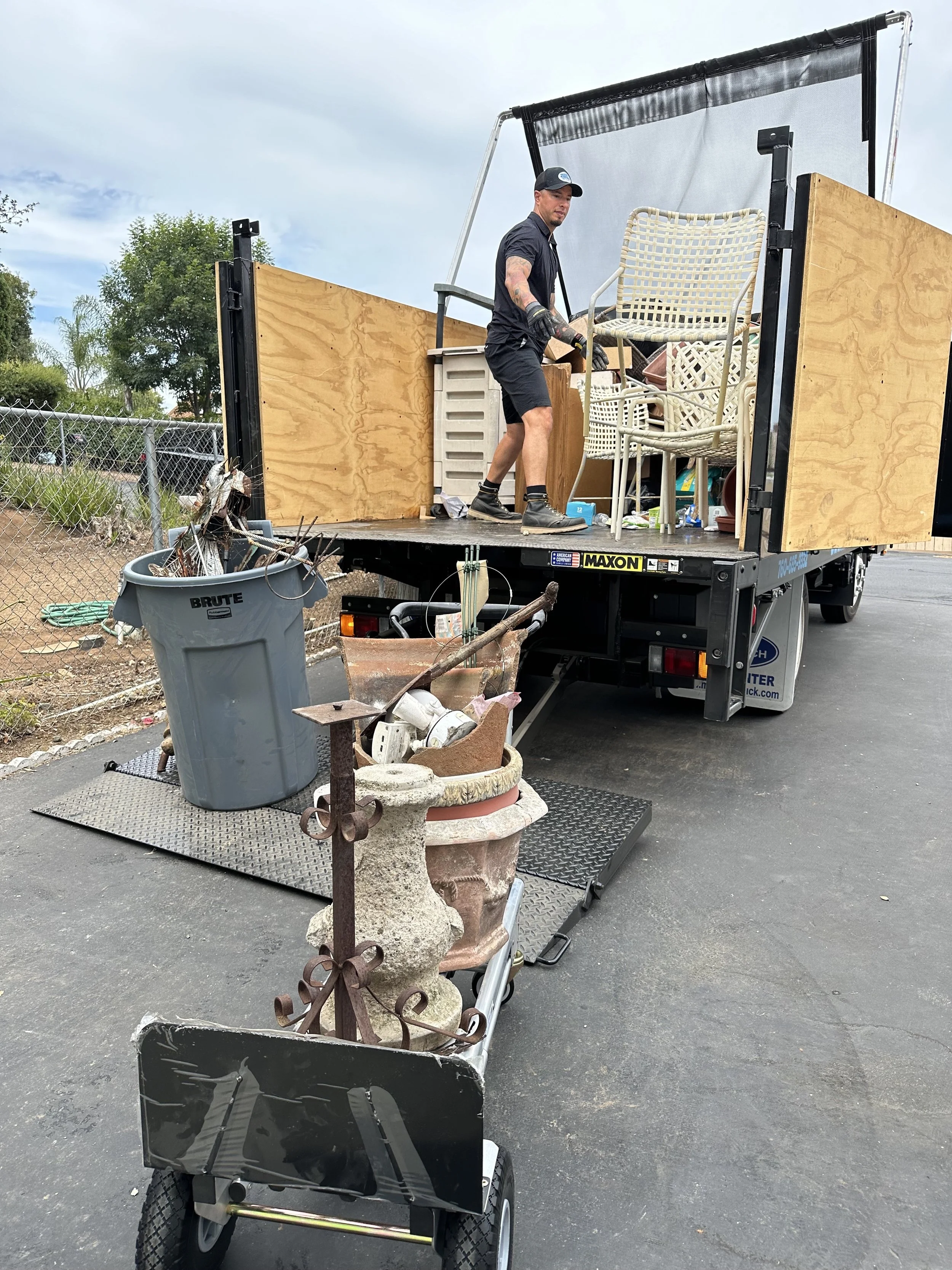 A man on a truck bed filled with furniture and household items, emptying a cart filled with garden pots and yard decorations.