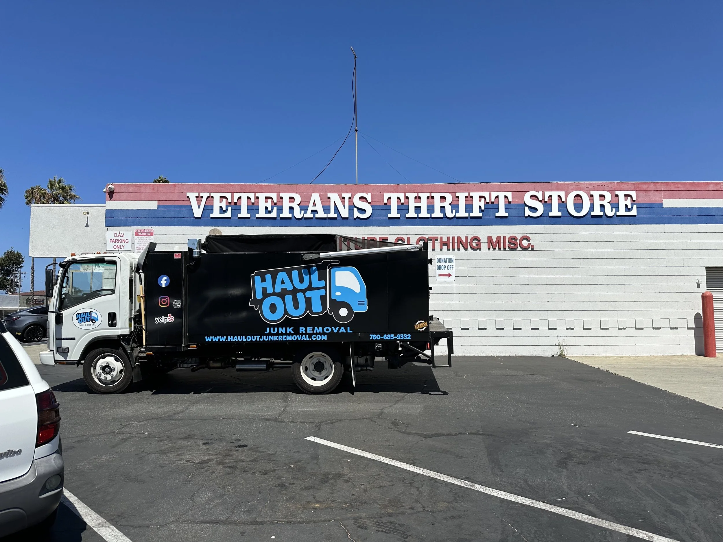 Parking lot in front of a Veterans Thrift Store with a donation drop-off sign, with a black truck labeled 'Haul Out Junk Removal' parked in the foreground, and palm trees against a clear blue sky in the background.