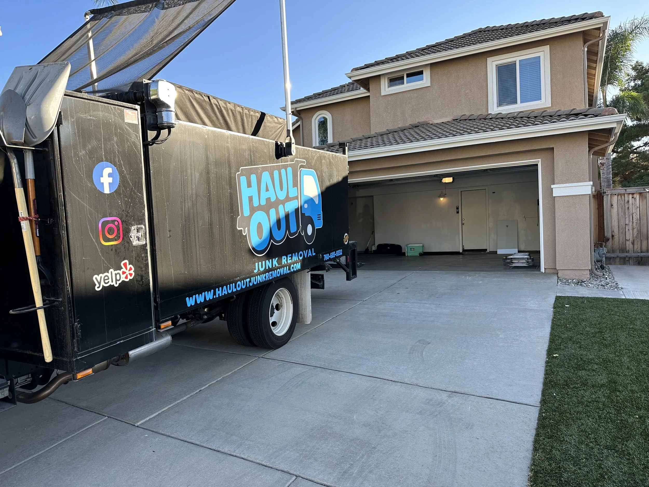 A black junk removal truck marked 'Haul Out' parked in front of an open garage in a residential neighborhood. The truck features icons for Facebook, Instagram, and Yelp, with a website URL.