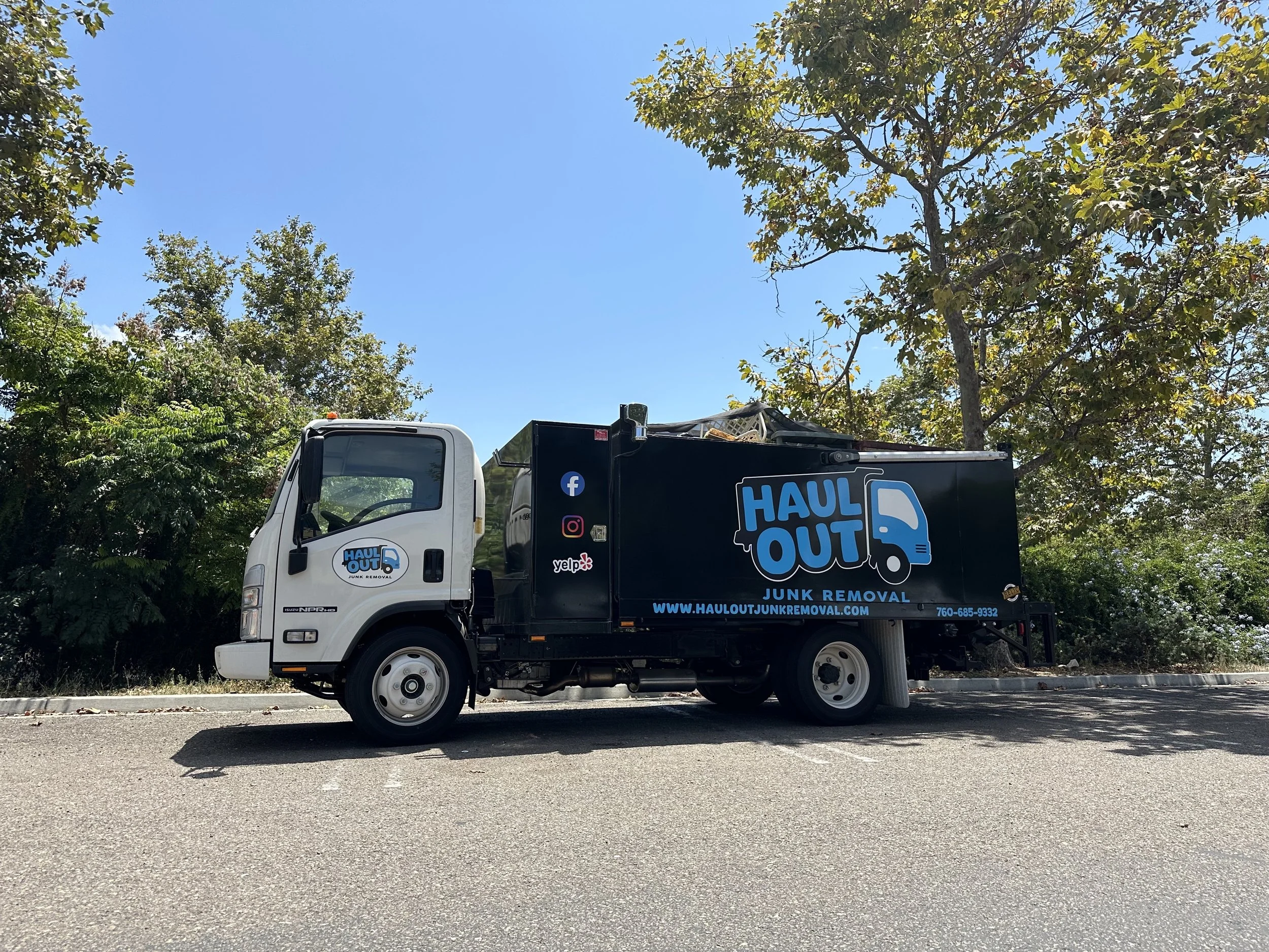 A white and black junk removal truck parked on the side of the road with trees and a blue sky in the background.