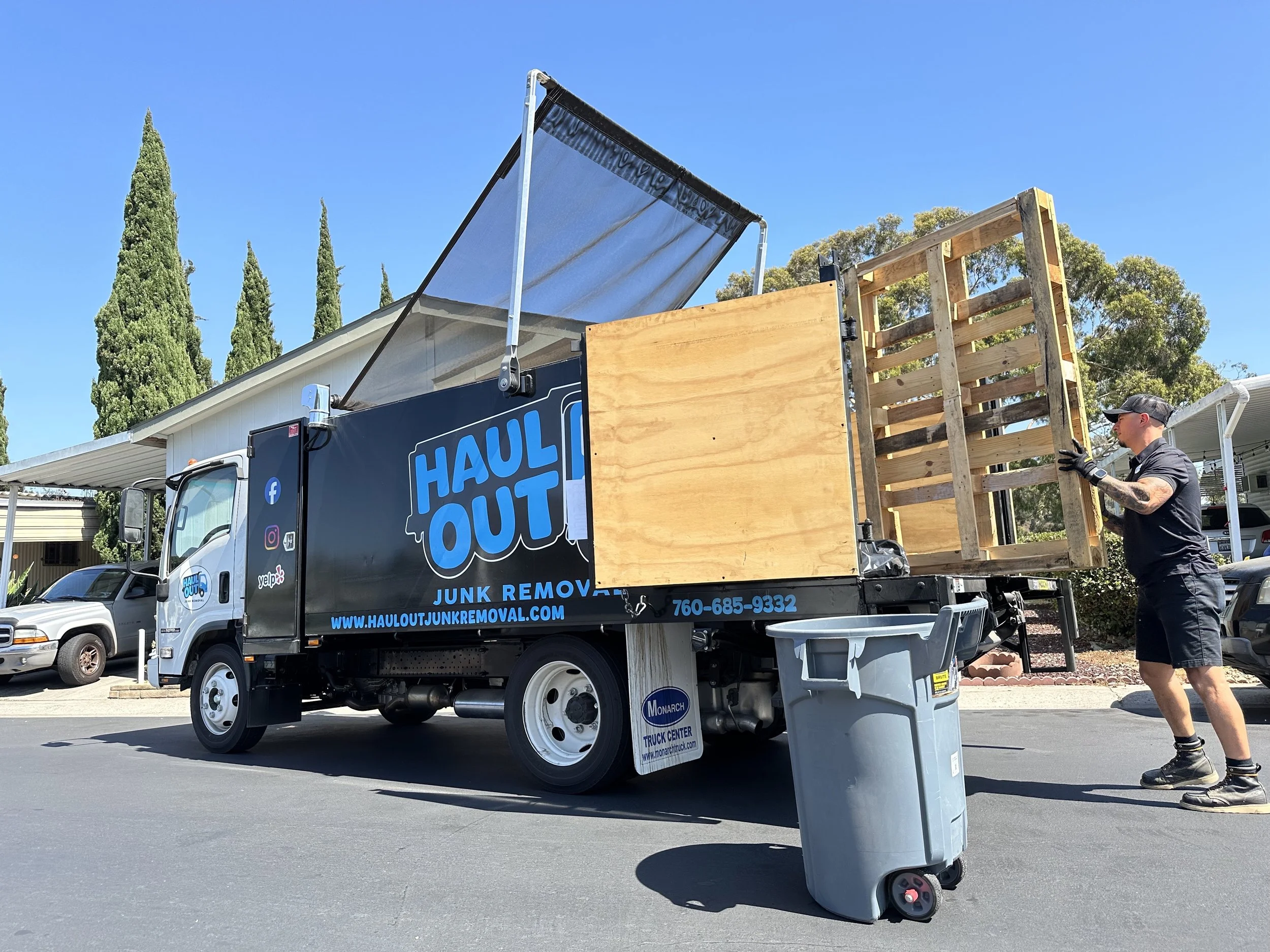A man loading a large wooden crate onto a flatbed truck labeled "Haul Out Junk Removal" on a sunny day with blue skies and tall trees in the background.