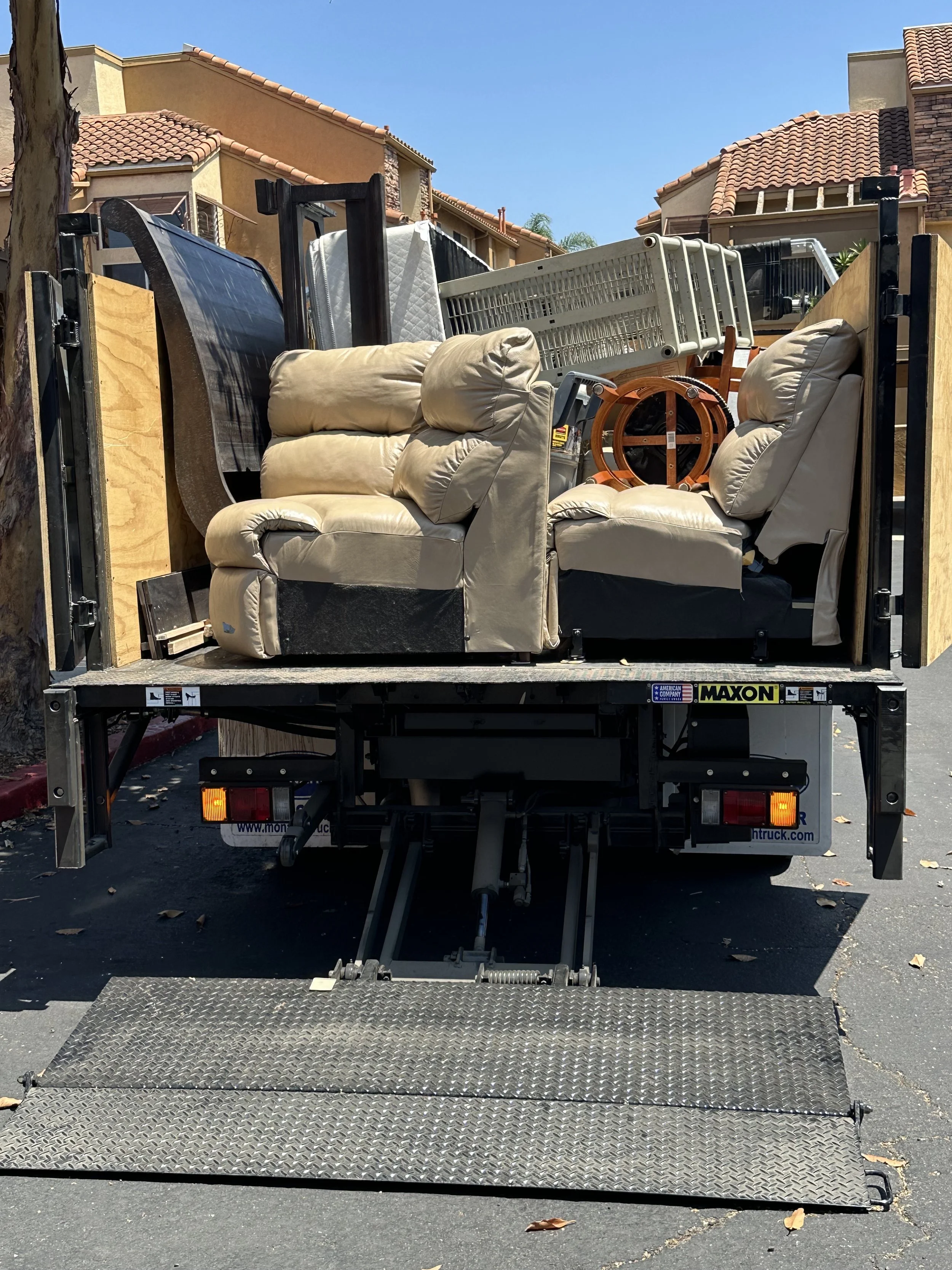 A moving truck with furniture and household items loaded inside, including beige couches, a wooden chair, and a baby crib, parked in a residential area with houses in the background.