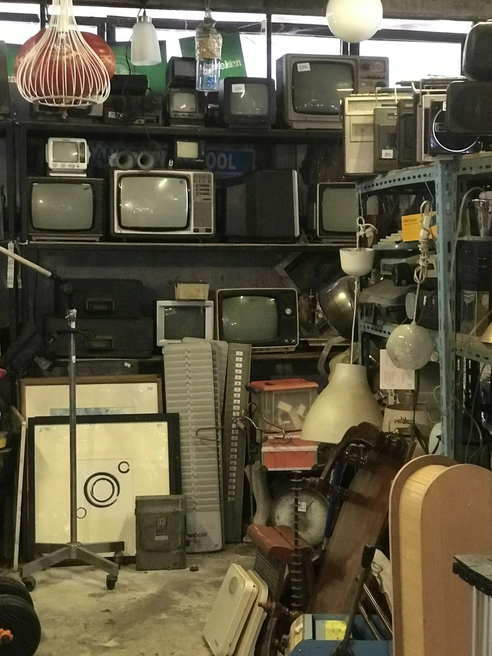 A cluttered storage room filled with various vintage televisions, lamps, and miscellaneous electronics and household items stacked on shelves and the floor.