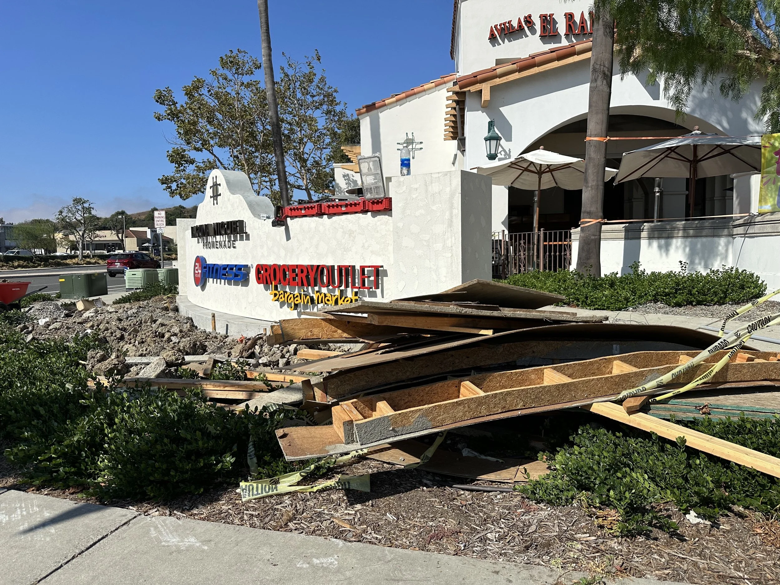 Construction debris, wooden planks, and caution tape in front of a white sign for a grocery outlet, fitness center, and a bargaain market, with a building labeled 'Avila's El Ranc' in the background.