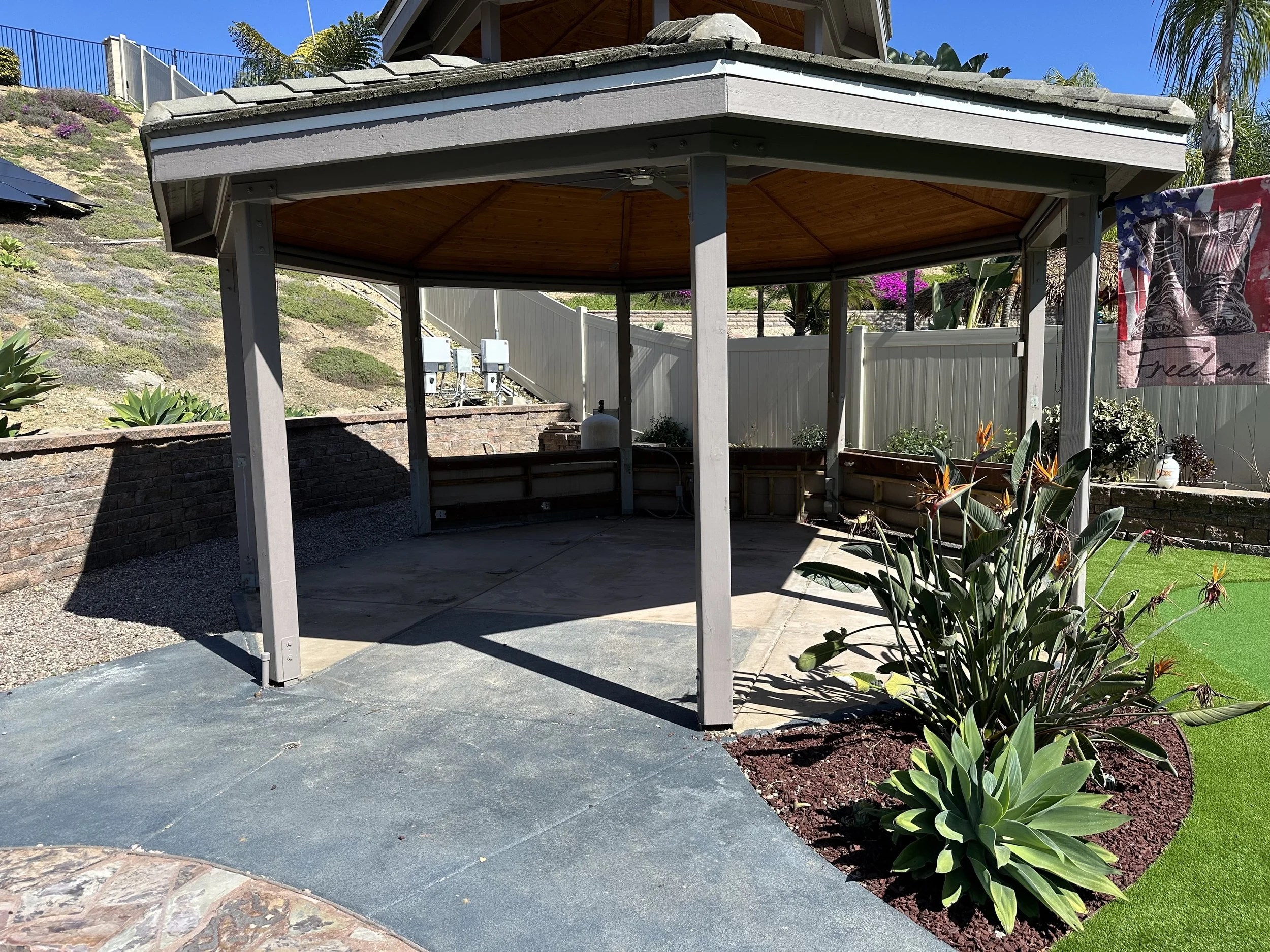 A backyard gazebo with a hexagonal wooden roof, supported by grey columns, on a concrete and stone patio, with plants and a colorful flag on the side.