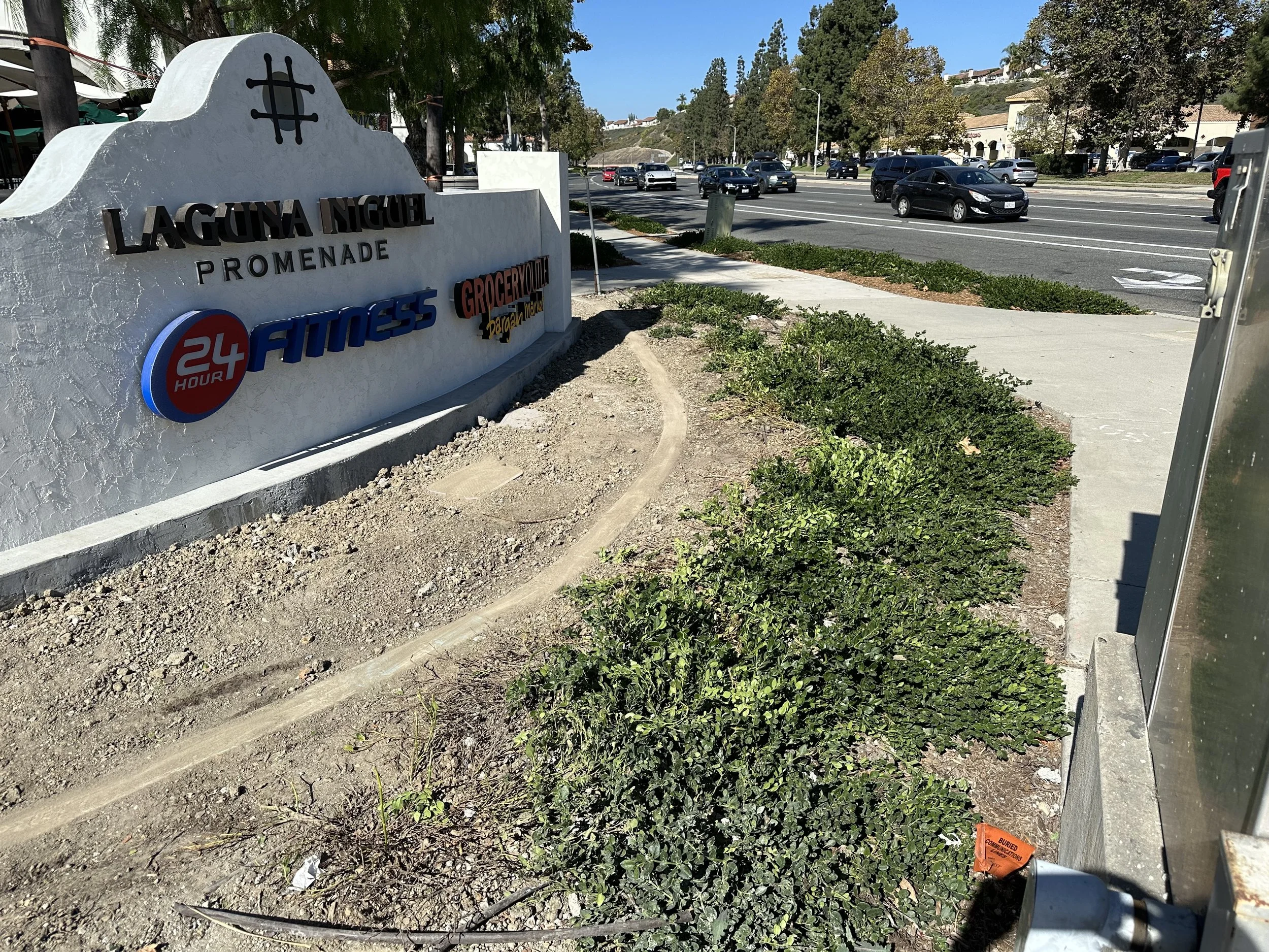 Sign for Laguna Niguel Promenade shopping center with logos for 24 Hour Fitness, Grocery Outlet, and other stores, along a sidewalk with cars on a street, trees, and a clear blue sky in the background.
